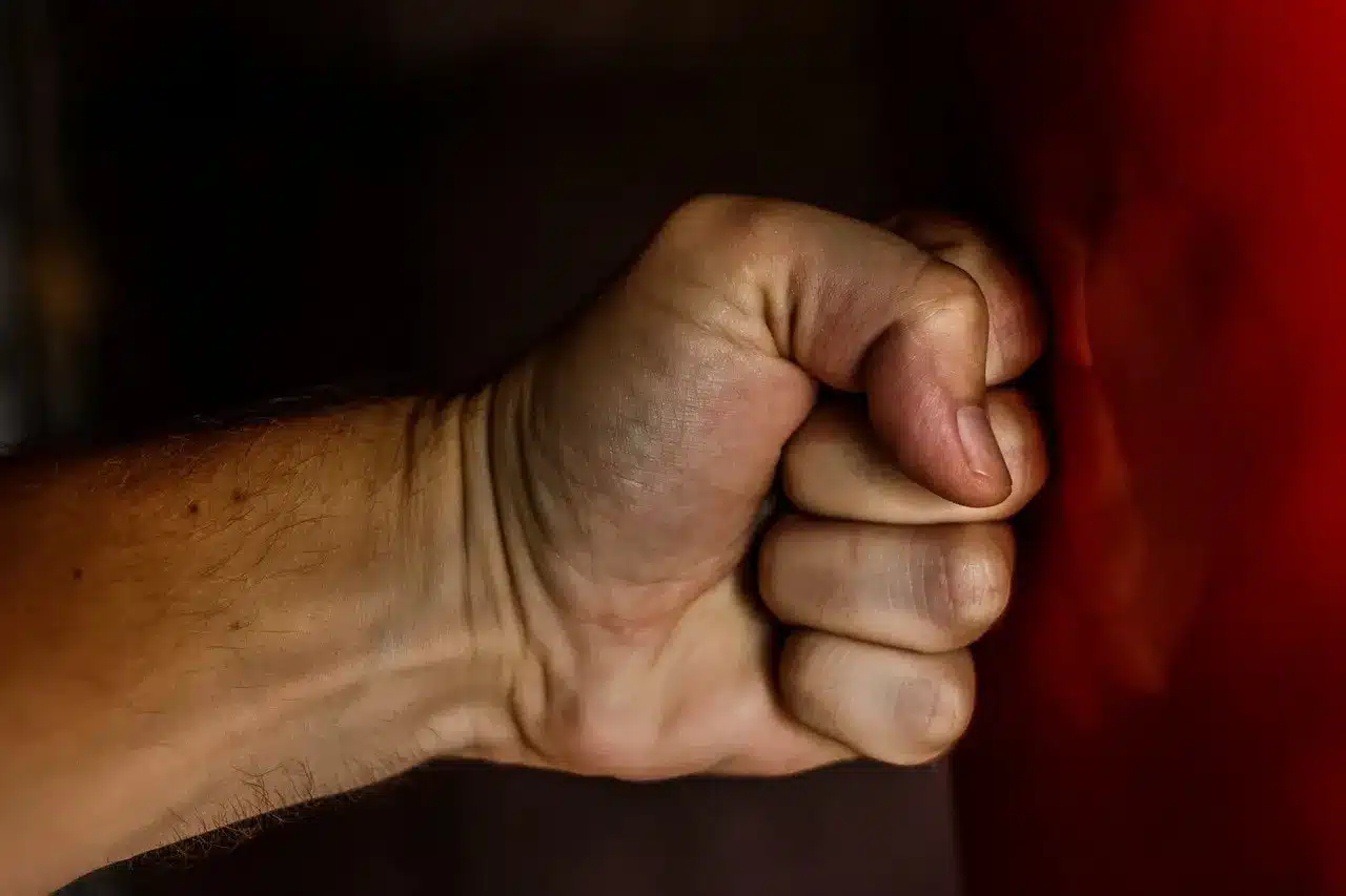 A close-up of a clenched fist with a light skin tone, positioned as if it is about to punch or is striking a red surface, against a dark background.