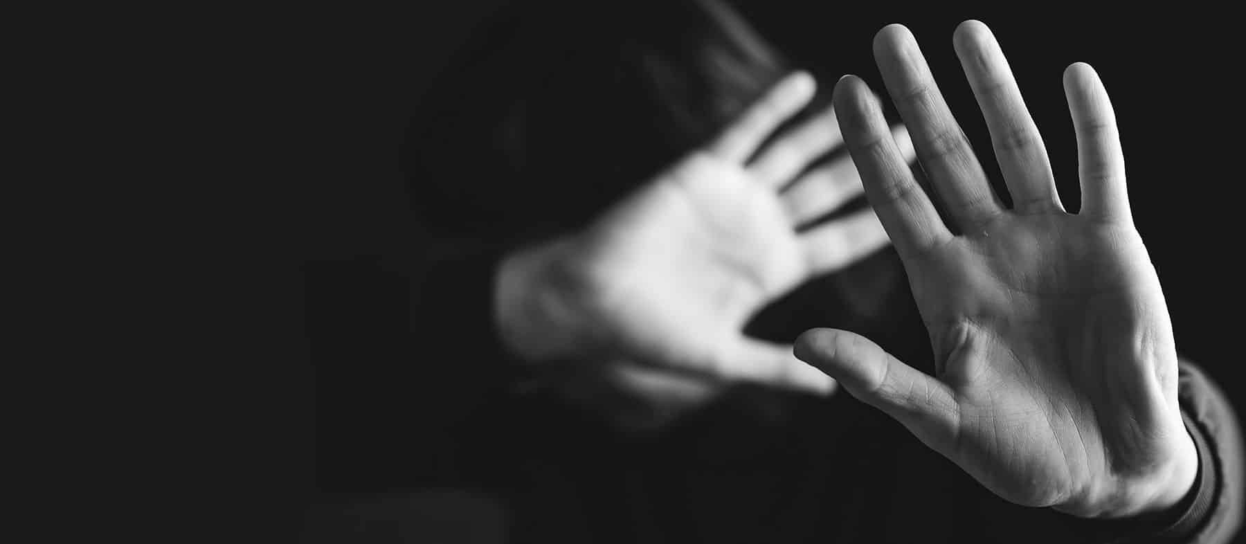 A person raising their hands in front of their face, as if shielding themselves or warding something off, in a black and white image with a dark background.