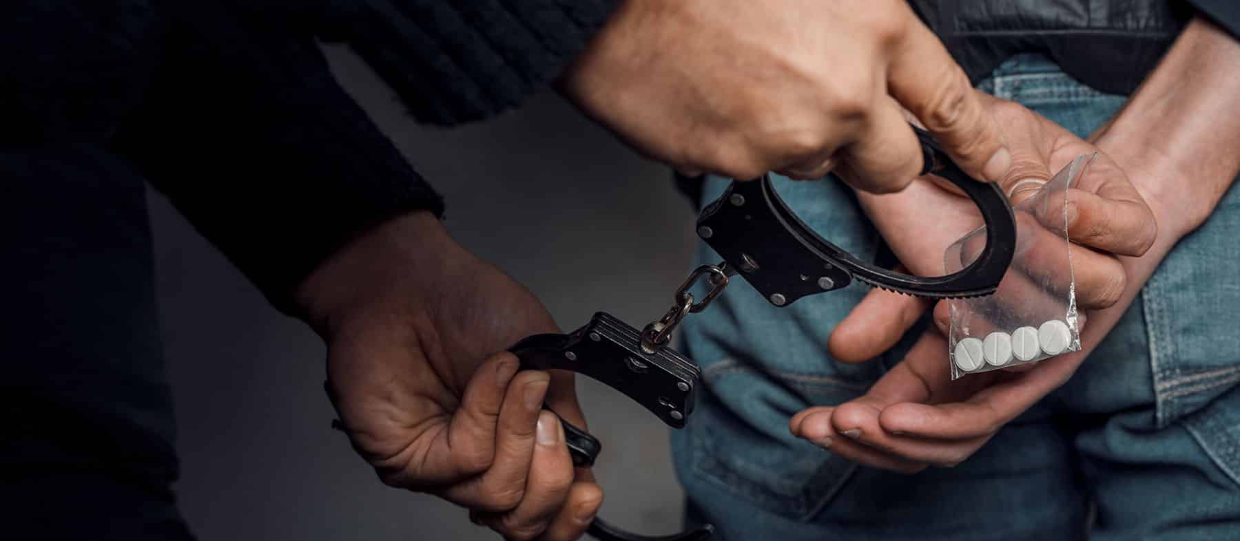 A person is being handcuffed while holding a small bag containing white pills behind their back, suggesting an arrest related to drugs.