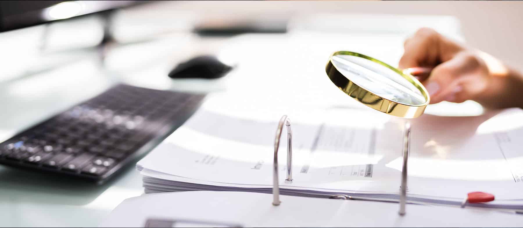 A person holds a magnifying glass over documents in a binder on a desk, with a computer keyboard visible in the background.