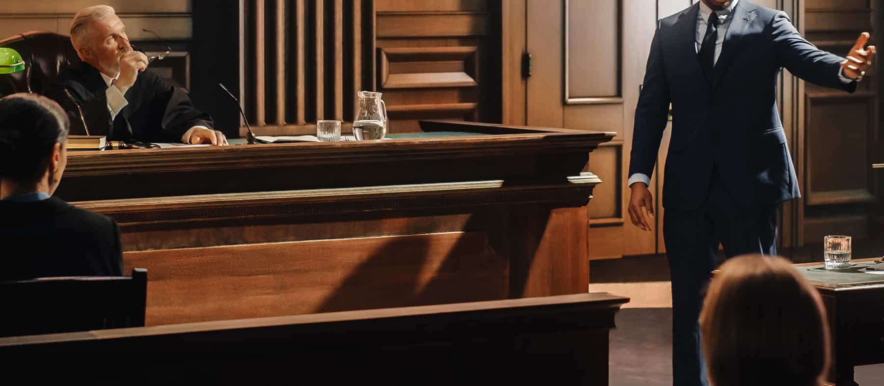 A lawyer in a blue suit speaks passionately in a courtroom, facing a judge seated at the bench, with several people watching from the audience.
