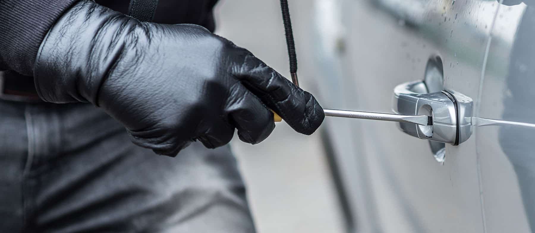 A person wearing black gloves uses a screwdriver to pry open the door lock of a silver car, suggesting an attempted car break-in or theft.