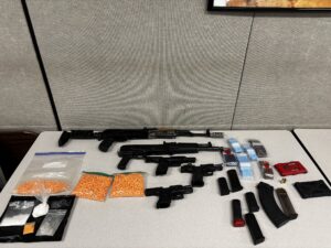 Two rifles, three handguns, several magazines, ammunition, and multiple plastic bags filled with orange and white pills laid out on a table.